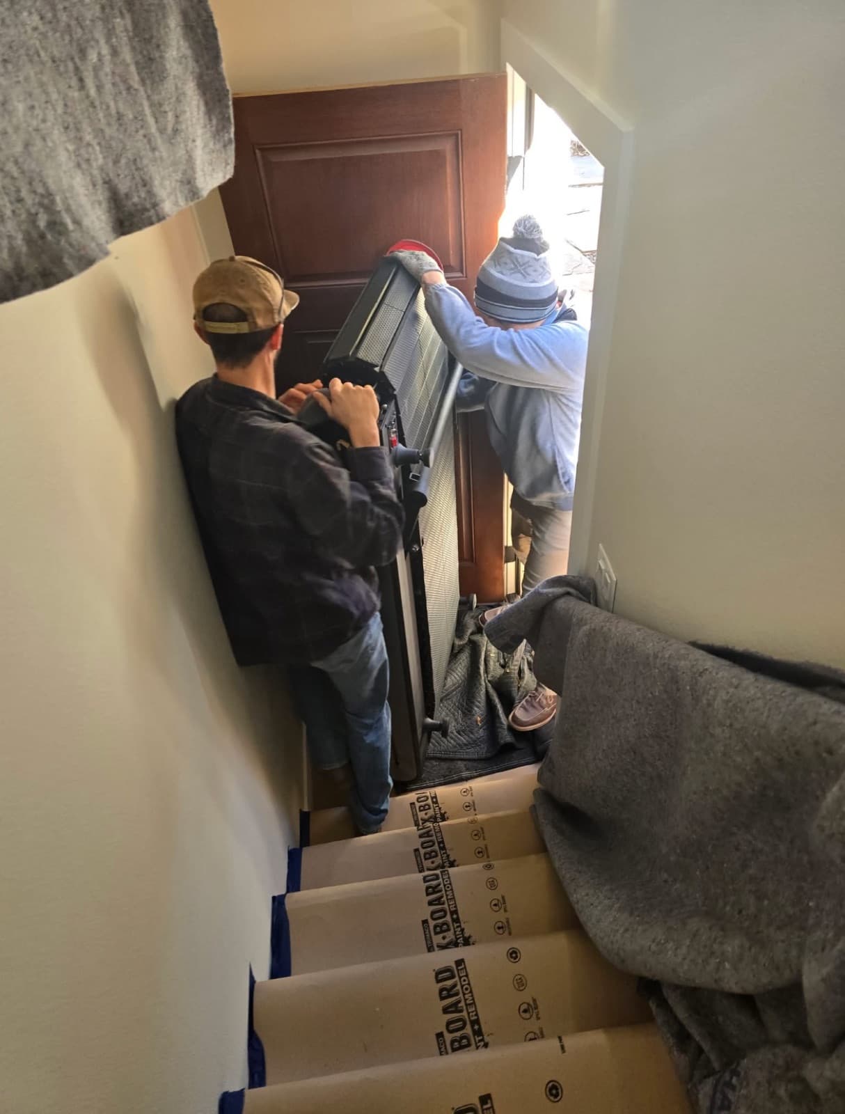 Two Heritage Moving crew members carrying a treadmill down a narrow staircase with cardboard floor protection